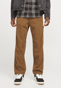 Brown corduroy pants with a straight cut, featuring vertical ribbing and front pockets. Paired with a black and white plaid shirt and black sneakers.