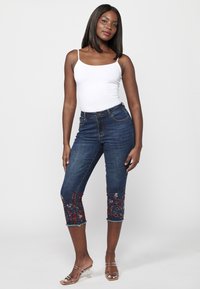 Blue denim capri jeans with floral embroidery at the hem, featuring a high waist and frayed edges. Paired with a white tank top.