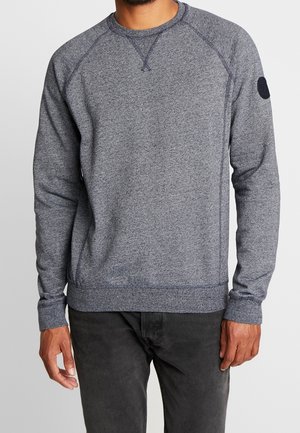 Sudadera gris con mangas largas, dobladillo y puños acanalados, mangas raglán y un parche circular en el hombro izquierdo. Textura suave.