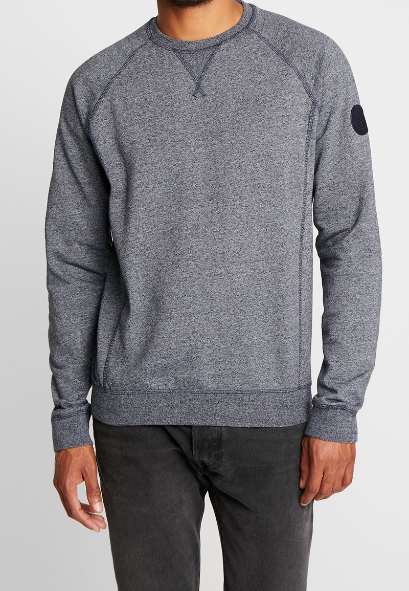 Sudadera gris con mangas largas, dobladillo y puños acanalados, mangas raglán y un parche circular en el hombro izquierdo. Textura suave.
