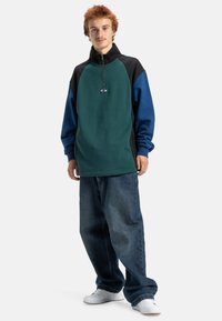 Pullover de fleece verde, preto e azul com meia zip; apresenta um logo no peito. Combinado com uns jeans de ganga azuis largos e ténis brancos.