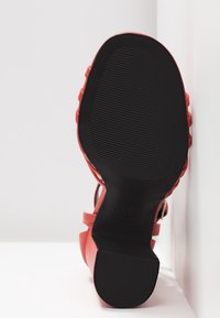 Semelle d'une sandale rouge avec une semelle en caoutchouc noir texturé et un petit talon bloc, présentée sur un fond blanc.