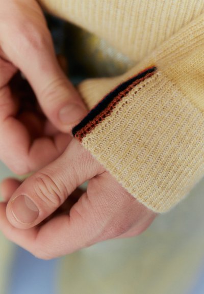 Sárga kötött pulóver mandzsetta bordázott textúrával, barna és sötétkék csíkkal. Kéz tartja a mandzsettát a csuklón.