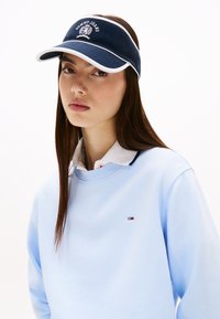 Vizuă navală cu bordură albă și logo, purtată cu un hanorac bleu deschis ce prezintă un logo mic. Textură netedă cu un design casual.