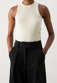 Top a maniche corte color crema con colletto alto, abbinato a pantaloni a vita alta neri con pieghe, mani nelle tasche.