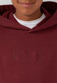Osoba ubrana w bordowy sweatshirt z wypukłym, teksturowanym napisem "SLVR" na klatce piersiowej oraz białą koszulę widoczną pod dekoltem.