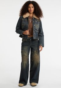 Giubbotto di denim con colletto in pelliccia sintetica tan, colore blu scuro, cinque tasche e chiusura a bottoni. Abbinato a jeans a gamba larga con dettagli testurizzati.