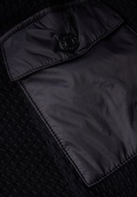 Svart texturerad tyg med en slät, glänsande svart klaff och en svart knapp. Designen har ett vävt mönster.