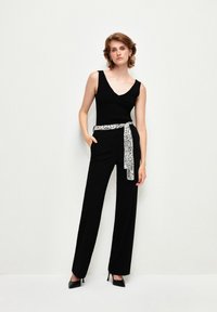 Zwarte mouwloze top met een v-hals, gecombineerd met zwarte wijde broek, geaccentueerd door een gestreepte witte riem. Het model staat zelfverzekerd.