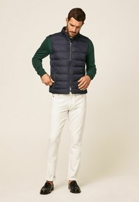 Marineblauer Steppweste mit hohem Kragen, Reißverschluss vorne und seitlichen Taschen, getragen über einem grünen Langarmshirt, kombiniert mit weißen Hosen und schwarzen Mokassins.