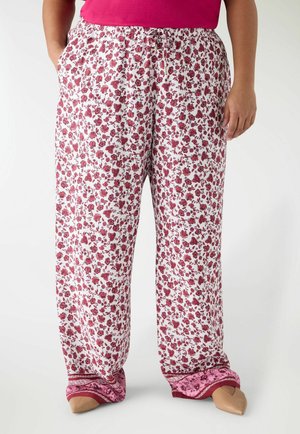 Donna che indossa pantaloni bianchi larghi con piccola stampa floreale rossa e polsini rosa decorati, abbinati a scarpe beige a punta.