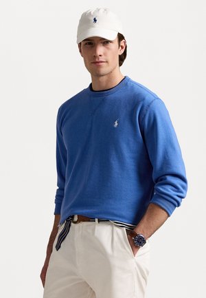 Blauer Pullover mit Rundhalsausschnitt und gesticktem Logo auf der Brust, kombiniert mit beigen pantalons und einer weißen Mütze. Am Handgelenk trägt er eine braune Uhr.