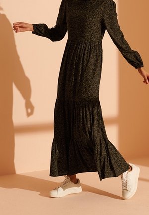 Person wearing a long, dark floral dress and white sneakers, walking on a peach-colored floor with a matching wall background.