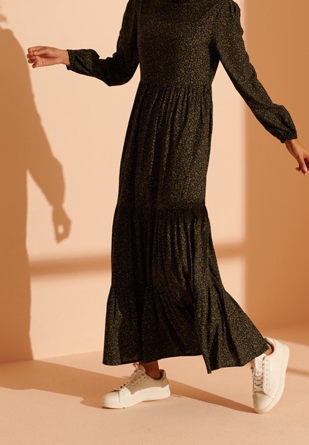 Personne portant une longue robe fleurie sombre et des baskets blanches, marchant sur un sol couleur pêche avec un mur assorti en arrière-plan.