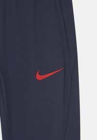 Marina blå träningsbyxor i slät tyg, med en utstickande röd Nike-logotyp på låret och sidofickor.