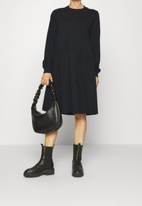 Mörkblå långärmad klänning med en plisserad kjoldel, i kombination med en svart texturerad handväska och kraftiga svarta ankelstövlar.