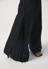 Pantaloni neri a gamba larga con dettaglio plissettato sul lato, tessuto morbido e texture liscia. L'orlo presenta uno strato trasparente sottile.