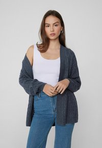 Dunkelgrau gestrickte Strickjacke mit offenem Design, gerippten Bündchen und überschnittener Schulter, getragen über einem weißen Tanktop und blauen Jeans.