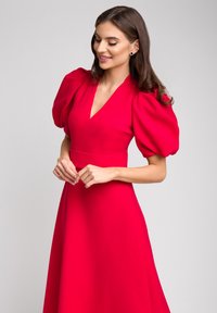 Czerwona sukienka do kolan z bufiastymi rękawami, dekoltem w serek oraz dopasowanym talią. Gładka tekstura i zwiewny dół tworzą elegancką sylwetkę.