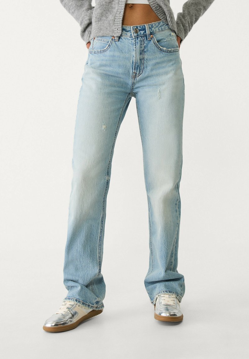 Personne portant un jean droit bleu clair légèrement usé et des baskets argentées, debout les mains dans les poches devant un fond uni.