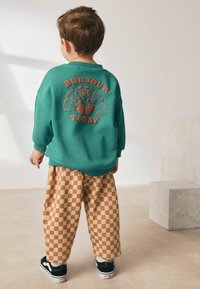 Felpa turchese con stampa "Bonjour Today", abbinata a pantaloni a quadri beige e marroni. Sneakers nere completano l'outfit.