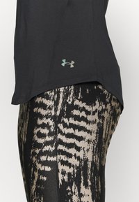 Top atletico nero con orlo più basso e logo verde Under Armour, abbinato a leggings con motivo astratto testurizzato nero e beige.