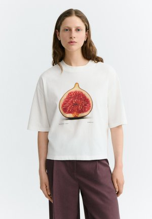 Jeune femme aux longs cheveux portant un t-shirt blanc avec un grand imprimé de figue, associé à un pantalon violet foncé, debout sur fond blanc.