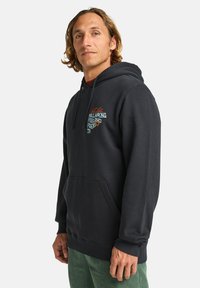 Man with wavy hair wearing a black hoodie featuring a colorful graphic, standing against a white background. Hands in pockets.