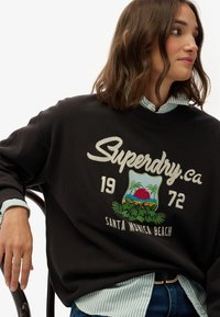 Czarny sweatshirt z haftowanym logo "Superdry" oraz wzorem plaży Santa Monica, z czerwonym słońcem i palmami. Nosi na sobie jasnoniebieską bluzkę w paski.
