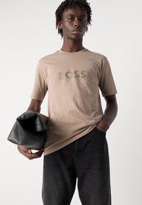 Beige bavlněné tričko s texturou s nápisem "BOSS" v zelených písmenech, spárované s tmavými džíny. Model drží černou koženou tašku.