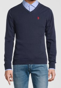 Navy V-hals trui met lange mouwen, geribbelde boorden en onderrand, met een klein rood logo op de borst, gedragen over een lichtblauw overhemd met kraag.