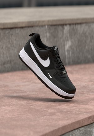 Pantof sport Nike Air Force 1 alb-negru atârnat deasupra unei suprafețe roz de beton, cu un fundal de piatră gri.
