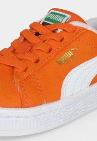 Πορτοκαλί σουέτ αθλητικό παπούτσι Puma με λευκή σόλα, λευκή πλαϊνή ρίγα, πορτοκαλί κορδόνια και πράσινη ετικέτα με το λογότυπο Puma στη γλώσσα.