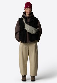 Gilet nero su una felpa marrone, pantaloni beige e stivali marroni. Indossa un berretto rosso e porta una borsa a tracolla grigia chiara. Pelle con lentiggini.
