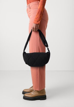 Black quilted handbag with a curved shape and a padded strap, held by a person wearing light pink pants and brown lace-up shoes.