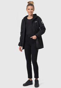 Veste imperméable noire avec capuche, dotée de poches zippées et d'accents de logo. Portée sur un haut noir et un jean foncé, accompagnée de baskets noires.