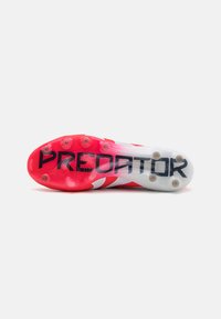 Semelle de chaussure de football rouge et blanche avec un motif texturé, comportant une marque "PREDATOR" noire et plusieurs crampons en caoutchouc pour l'adhérence.