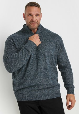 Homme aux cheveux courts portant un pull zippé en maille gris foncé, tenant le col d'une main sur un fond blanc uni.