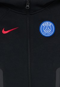 Black zip-up jacket with red Nike logo on left and blue circular Paris Saint-Germain emblem on right chest.