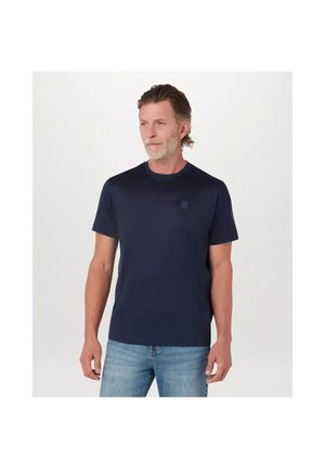 Man met kort grijs haar en baard, draagt een effen marineblauw t-shirt en lichtblauwe spijkerbroek, staand tegen een witte achtergrond.