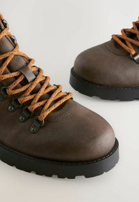 Brune lædervandre støvler med orange snørebånd og sorte robuste såler på en lys baggrund.