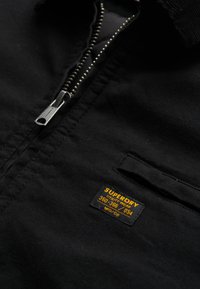 Veste noire zippée en tissu texturé, ornée d'une petite étiquette orange portant la mention "SUPERDRY" et d'une poche sur la poitrine.