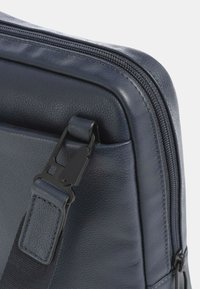 Borsa in pelle blu all'angolo con zip, cuciture visibili e una clip nera in metallo sulla tracolla, caratterizzata da una texture liscia e una forma strutturata.