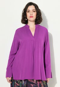 Camicia a maniche lunghe viola con colletto a punte, texture liscia e vestibilità ampia, abbinata a una gonna plissettata colorata.