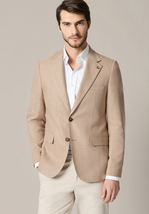 Mężczyzna w beżowej marynarce na białej koszuli i jasnych spodniach, stojący na neutralnym tle, ręce swobodnie opuszczone wzdłuż ciała.