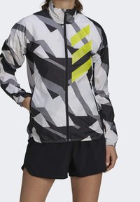 Let vindjakke med geometrisk mønster i sort, hvid og grå med en fremtrædende gul accent. Lynlås hele vejen, høj krave.