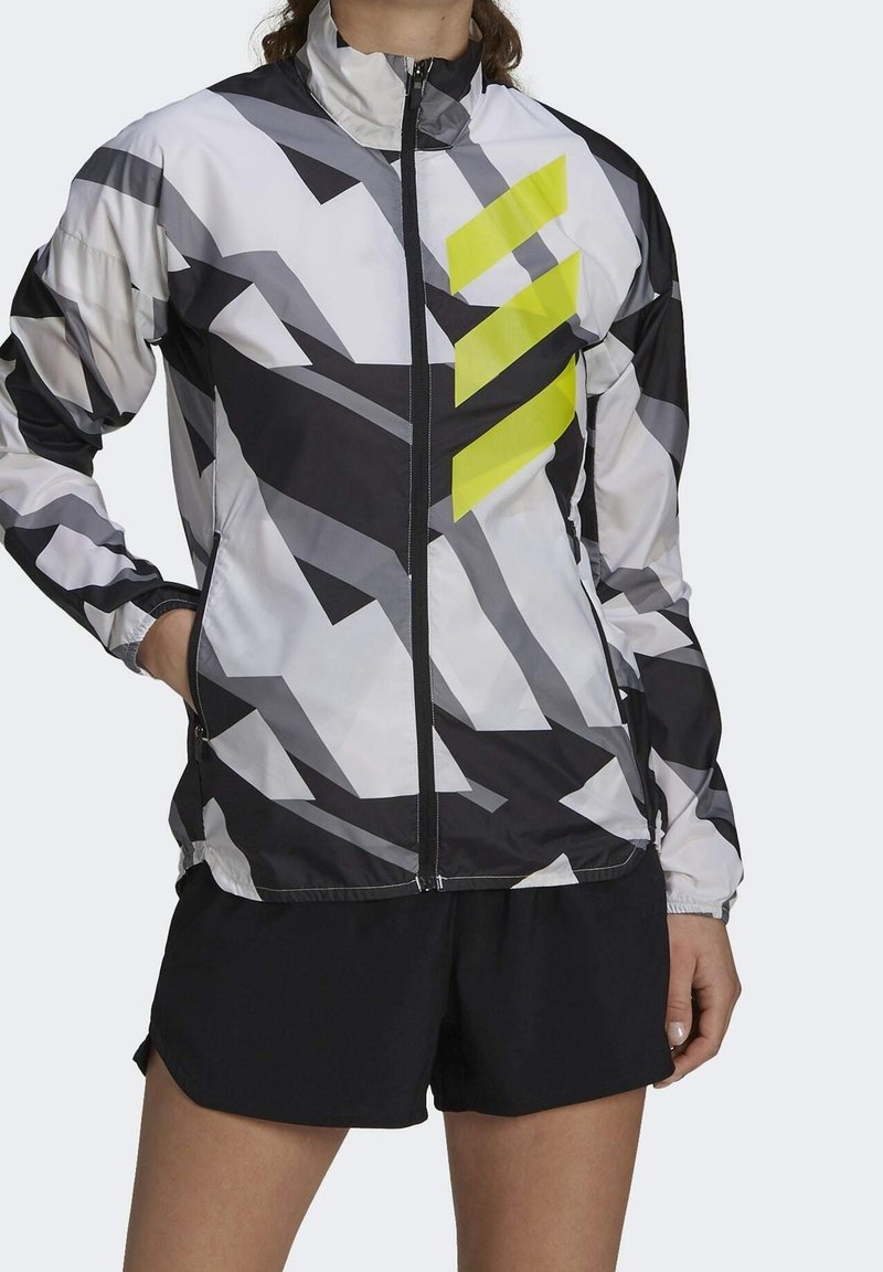 Let vindjakke med geometrisk mønster i sort, hvid og grå med en fremtrædende gul accent. Lynlås hele vejen, høj krave.