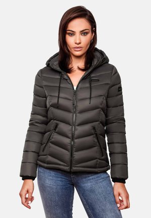 Femme portant une veste noire matelassée à capuche avec des poches zippées, associée à un jean bleu, debout devant un fond clair uni.