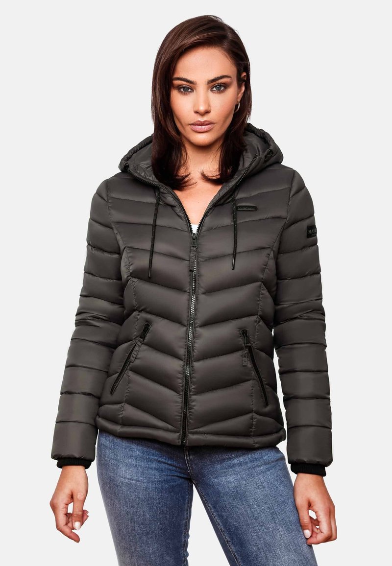 Femme portant une veste noire matelassée à capuche avec des poches zippées, associée à un jean bleu, debout devant un fond clair uni.