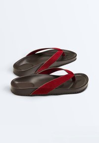 Chaussons marron foncé avec des brides en daim rouge en forme de Y, dotés de semelles intérieures moulées et de semelles texturées, présentés sur fond blanc.